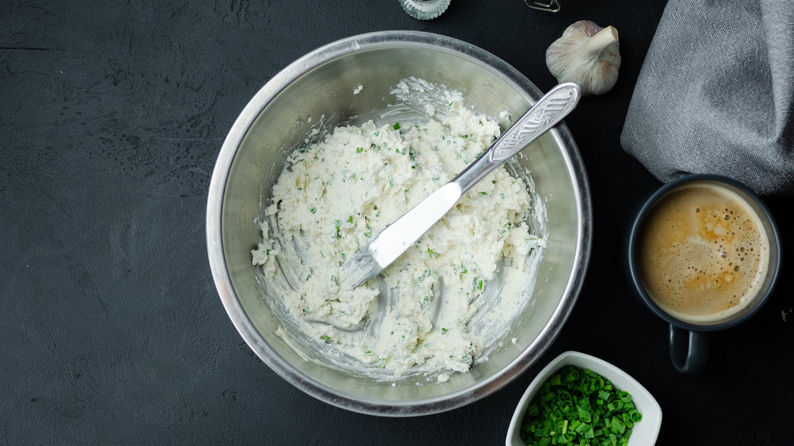 Top view of a stainless steel bowl with creamy herb cheese spread, knife and coffee.