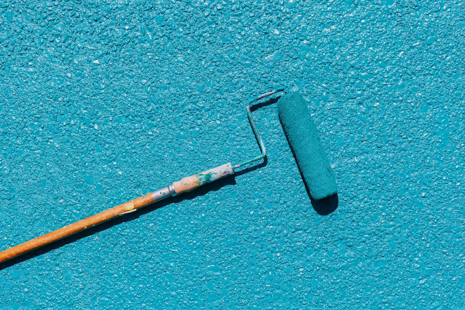A close-up of a blue paint roller applying paint on a textured wall surface, captured in bright sunlight.