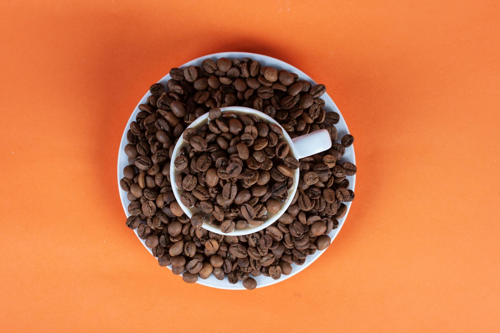 A cup filled with coffee beans on a saucer against an orange background viewed from above.