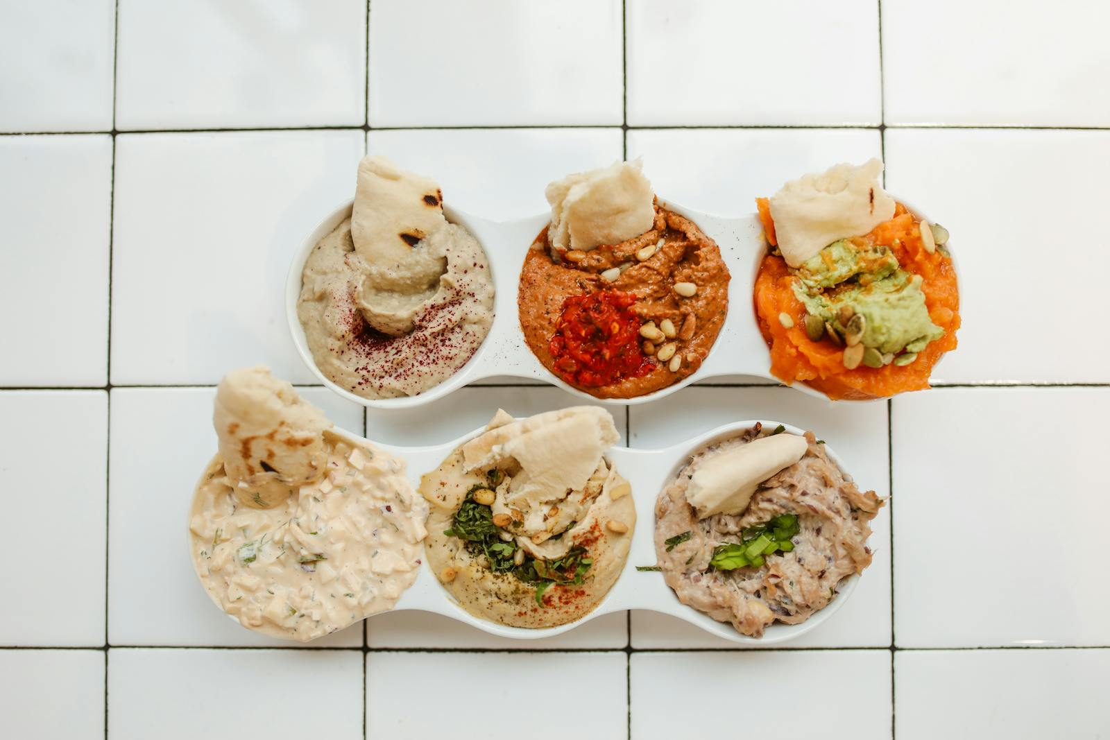 Overhead view of various dips with garnishes and bread on a white tiled background.