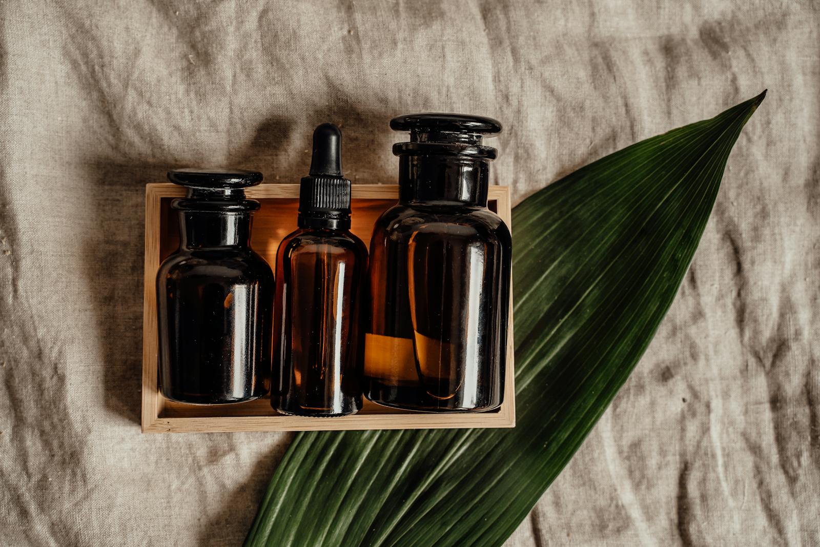 Collection of brown glass bottles in a wooden box with a green leaf on burlap fabric, perfect for organic skincare themes.